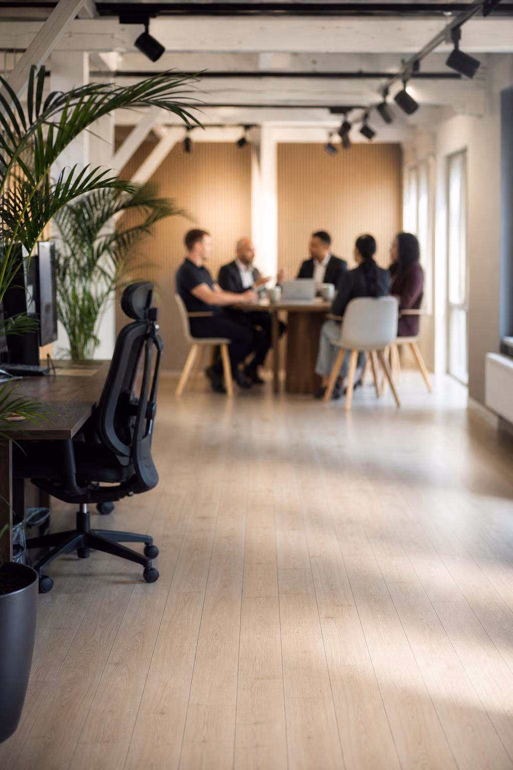 Team overlegt rond een houten tafel in een lichte loftkantoorruimte met planten en een bureau op de voorgrond.