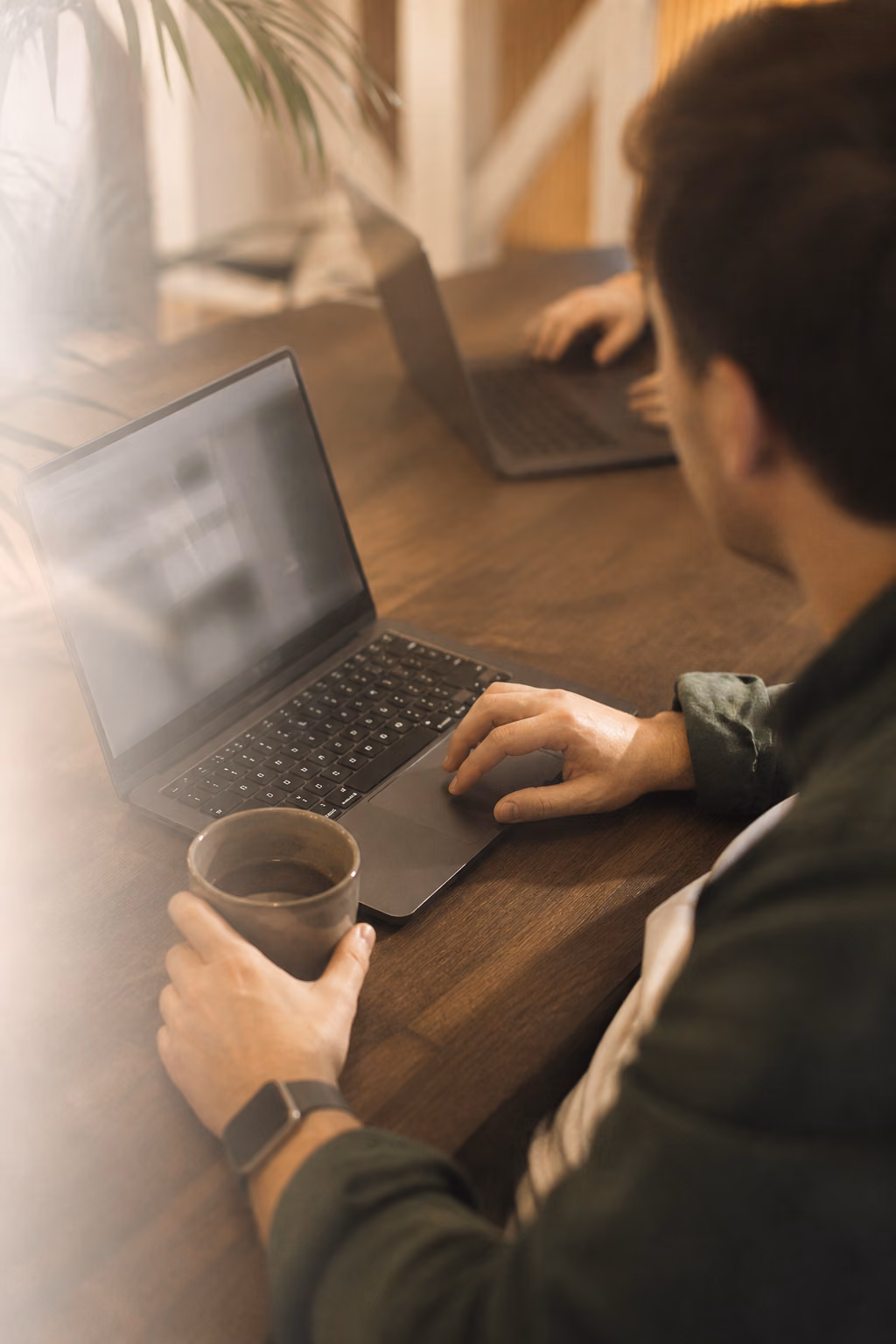 Man werkt op een MacBook aan een walnoten bureau met een kop koffie, met op de achtergrond een collega aan een tweede laptop.