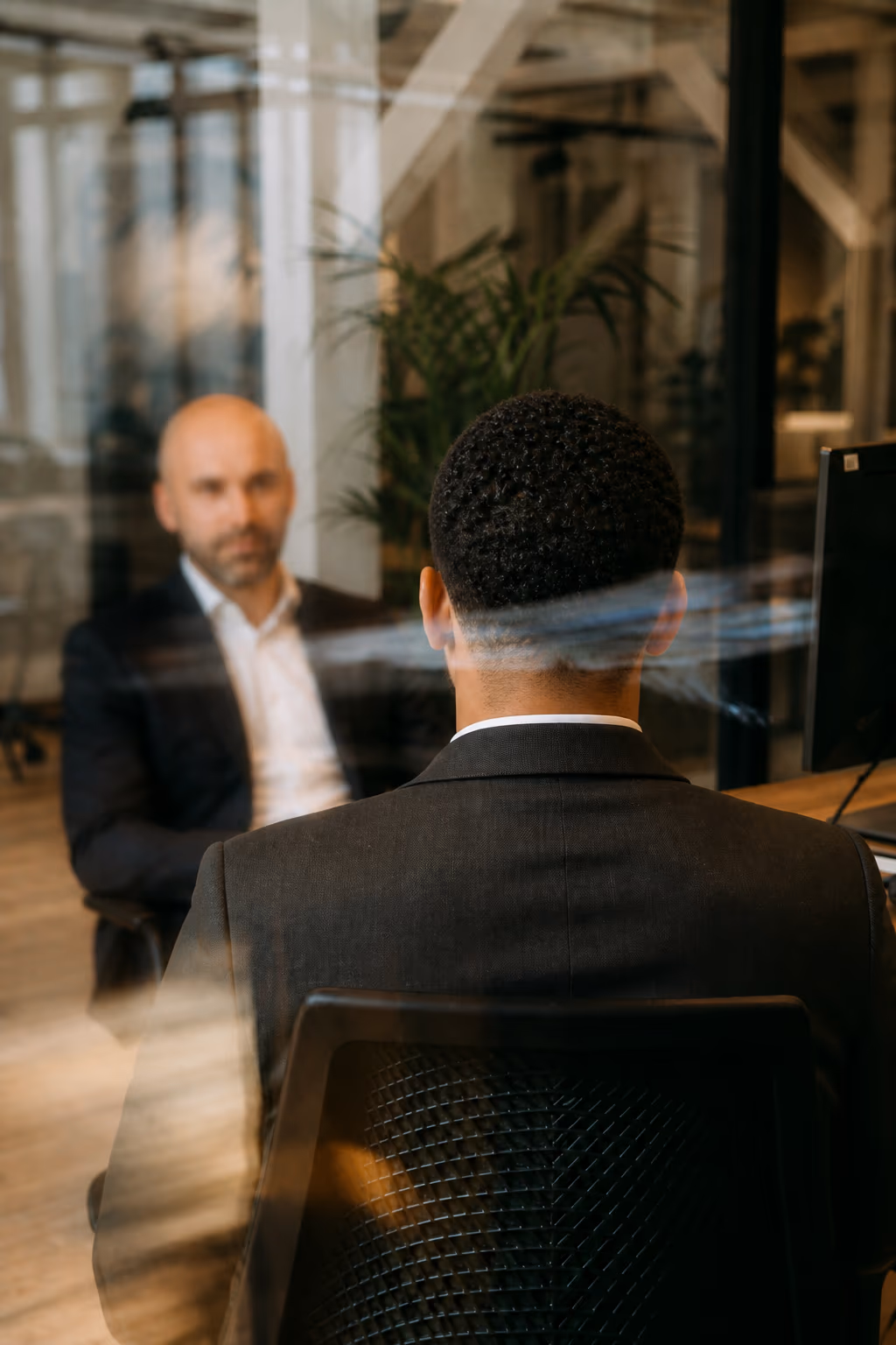 Twee mannen in pak overleggen aan een bureau in een kantoor, gefotografeerd door een glazen wand.