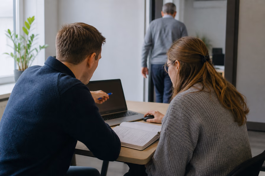 Twee collega's zitten samen aan een laptop, de een wijst met een pen iets aan terwijl de ander meeschrijft in een notitieboek.